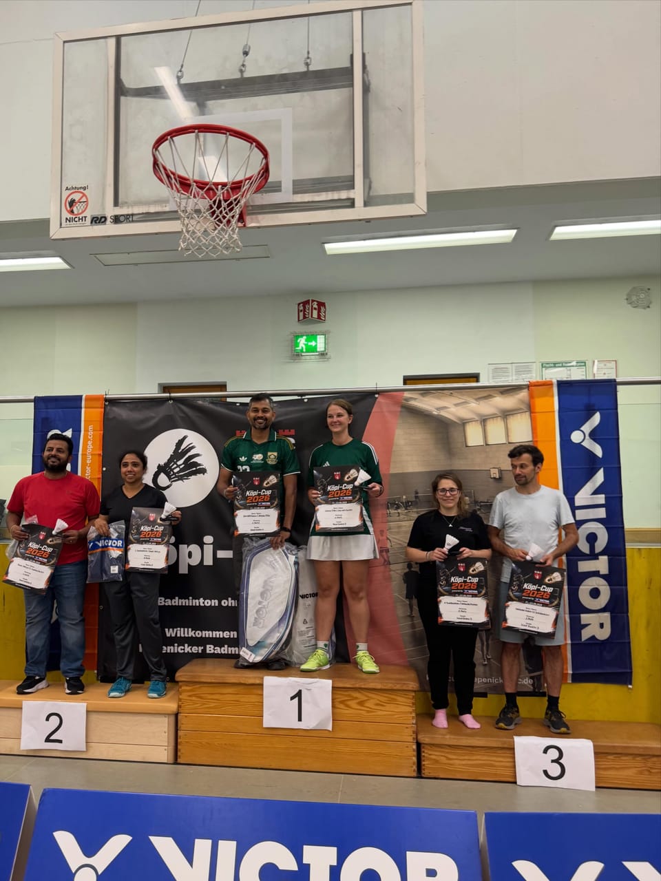 Sechs Personen mit Urkunden, Federbällen und einer Schlägertasche auf dem dreistufigen Siegerpodest in der Halle.