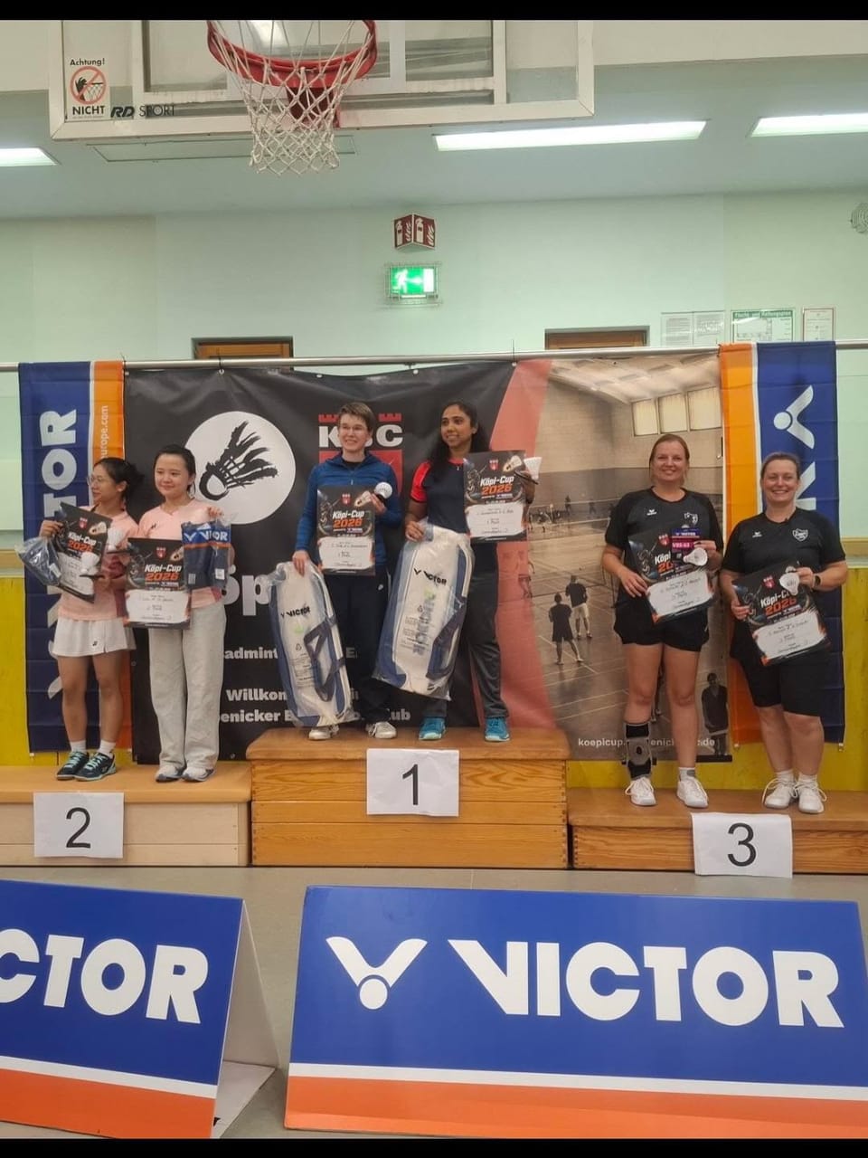 Sechs Spielerinnen mit Urkunden und Preisen auf dem Siegertreppchen vor dem Turnierbanner in der Halle.