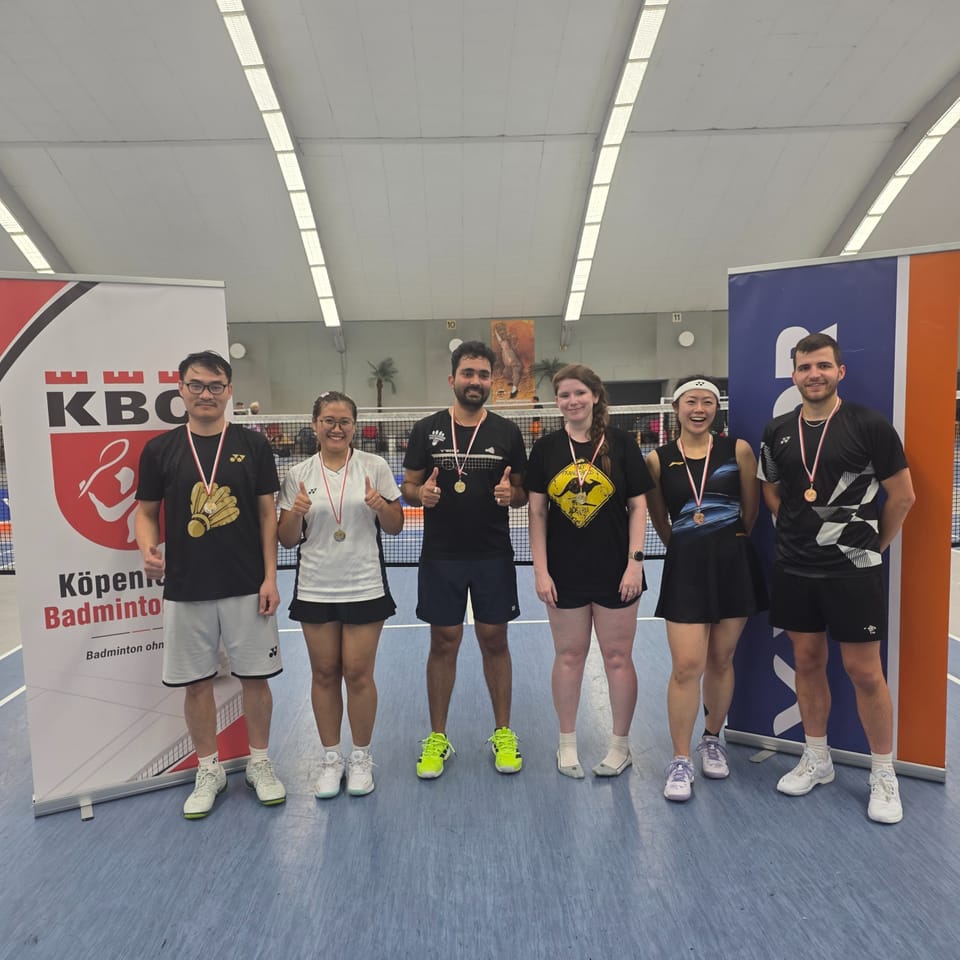 Sechs Spielerinnen und Spieler mit Medaillen zeigen lachend den Daumen nach oben, im Hintergrund KBC-Rollup und Badmintonfelder.